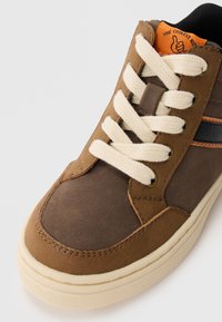 Tenis brown cu finisaj din piele de căprioară, șireturi crem și talpă din cauciuc texturat. Prezintă un accent cu dungi negre și portocalii pe lateral.