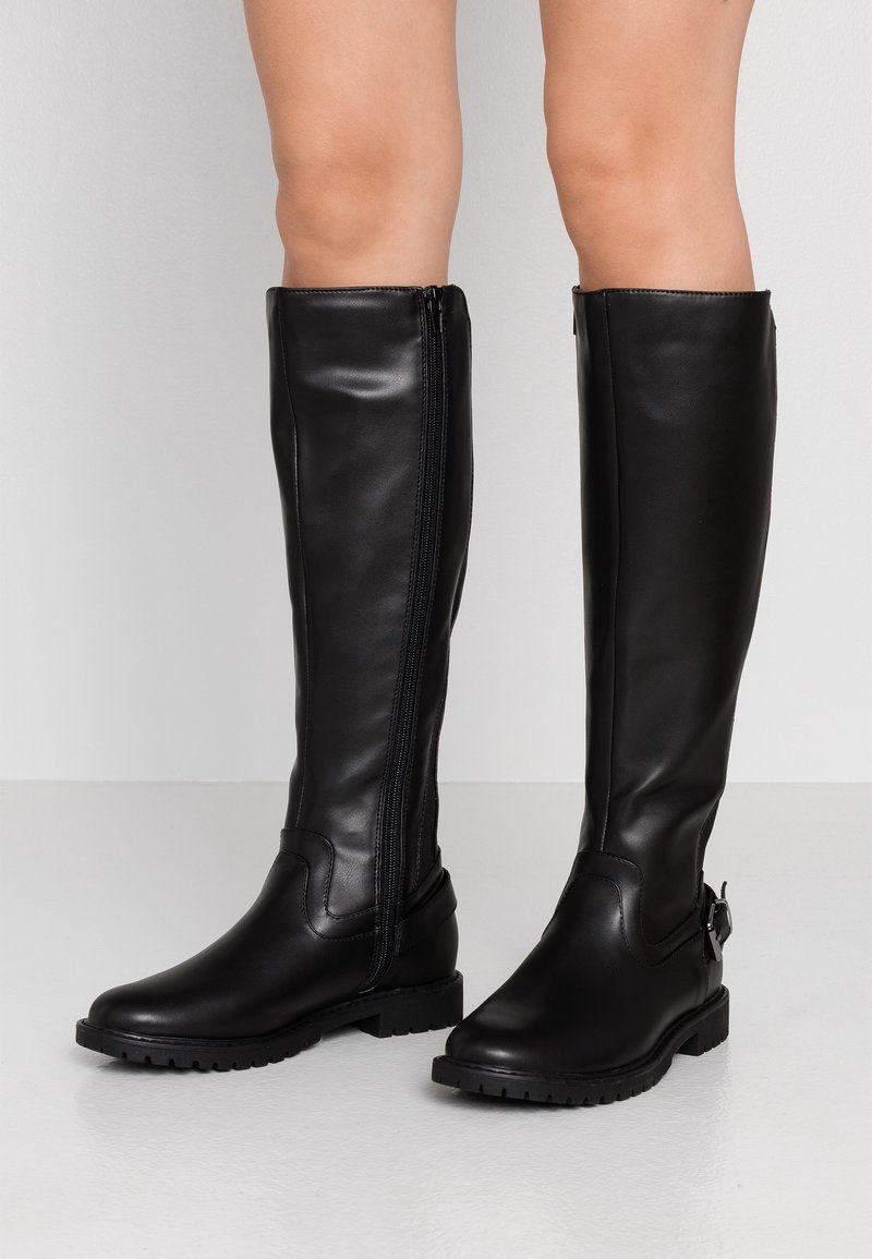 Ben klædt i knæhøje sorte læderstøvler med lynlås på siden og lave blokhæle, står på et glat hvidt gulv mod en hvid væg.