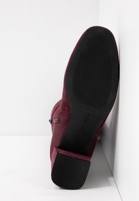 Semelle d'une botte bordeaux avec une semelle extérieure en caoutchouc noir texturée et un petit talon bloc reposant contre un mur d'angle blanc.