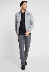 Siva flis jakna z visokim ovratnikom in zadrganim žepom na prsih, kombinirana s črno majico in sivimi hlačami, nošena s temnimi supergami.