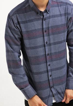 Mężczyzna w koszuli z długim rękawem zapinanej na guziki, z poziomymi paskami w odcieniach ciemnoszarym, niebieskim i stonowanym czerwonym, poprawiający mankiet na prawej ręce.