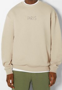 Beige sweatshirt met een ronde hals en geribbelde manchetten, met "PARIS" geborduurd in een lichtbruine lettertype aan de voorkant. Gedragen over een wit shirt.