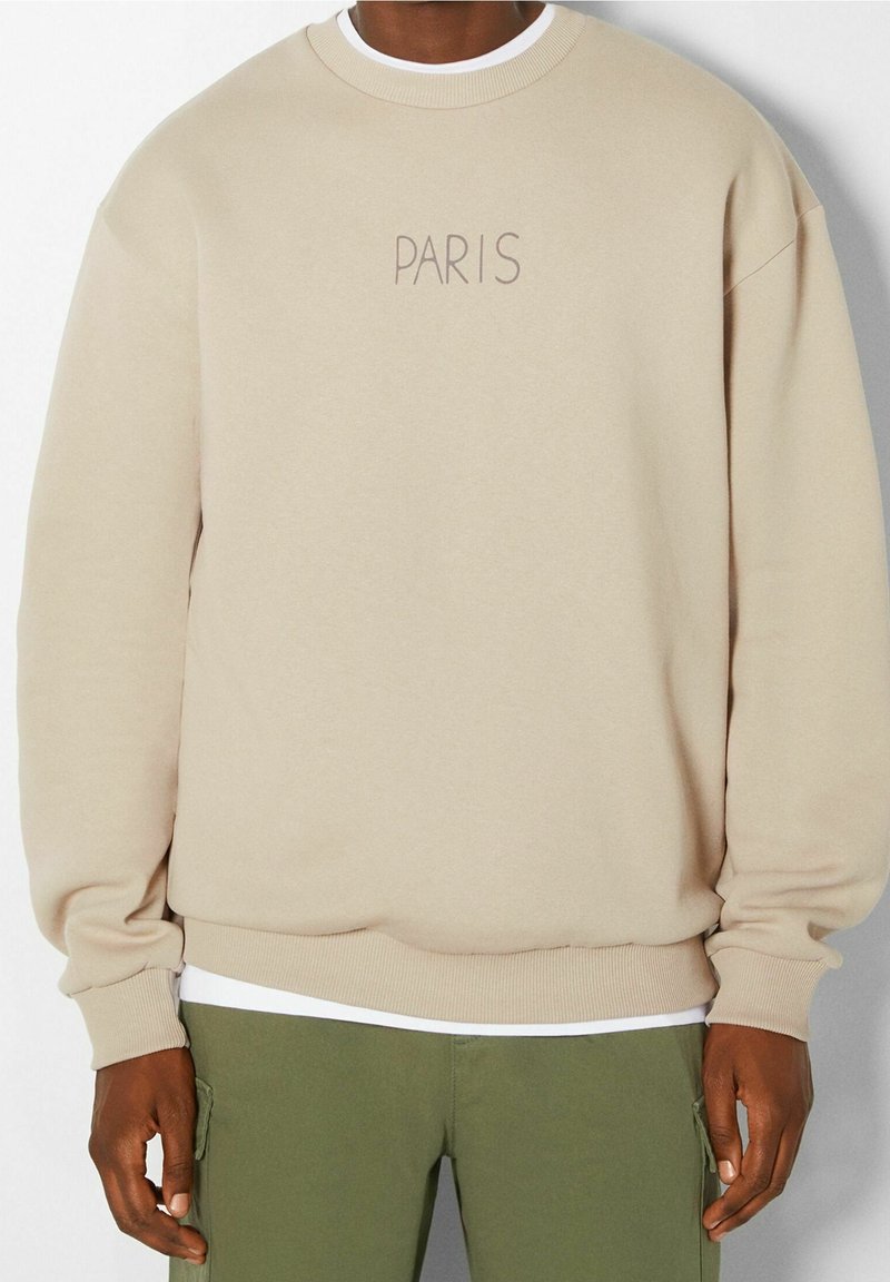 Beige sweatshirt met een ronde hals en geribbelde manchetten, met "PARIS" geborduurd in een lichtbruine lettertype aan de voorkant. Gedragen over een wit shirt.
