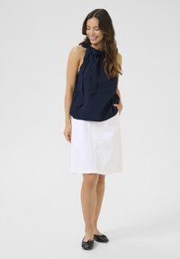 Haut sans manches bleu marine avec un col à nouer, associé à une jupe blanche arrivant aux genoux. Des chaussures plates noires complètent la tenue. Textures lisses et design simple.