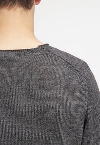 Gros plan de l'arrière d'une personne portant un pull en laine gris foncé avec des détails visibles de points sur l'épaule et l'encolure.