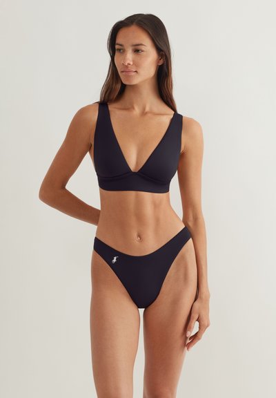 Γυναίκα που φοράει μαύρο μπικίνι με τοπ βαθύ V και σλιπ με ψηλή γραμμή, με μικρό λευκό λογότυπο στην αριστερή πλευρά.