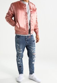 Rosa Satin-Bomberjacke, graues T-Shirt, ausgewaschene blaue Jeans und weiße Sneaker. Die Jacke hat gerippte Ärmelbündchen; die Jeans weisen verschiedene Risse auf.