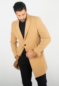 Manteau en mélange de laine couleur camel avec une fermeture à un bouton, des revers crantés et des poches latérales. Présente une coupe ajustée et une texture lisse.