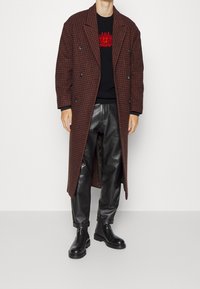 Brunrutigt dubbelknäppt kapp med en svart tröja med röd broderi, kombinerad med svarta läderbyxor och kraftiga svarta stövlar.