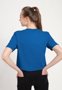 Niebieska koszulka z krótkim rękawem i okrągłym dekoltem, wykonana z gładkiej tkaniny, widziana od tyłu, noszona z czarnymi spodniami.