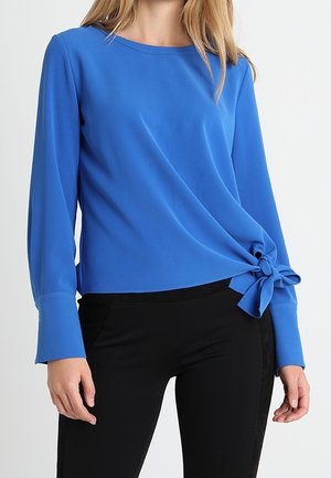 Femme portant un haut bleu à manches longues avec un nœud noué sur le côté droit et un pantalon noir sur un fond uni.