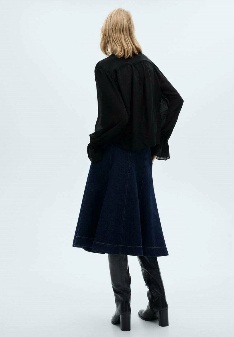 Femme blonde debout, dos à la caméra, portant un chemisier noir ample, une jupe midi bleu foncé, et des bottes noires hautes à talons.