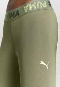 Legginsy Puma w oliwkowym kolorze z szerokim, elastycznym pasem, na którym znajduje się napis "PUMA" w kolorze żółtym. Biały logo na udzie, wykonane z gładkiego, elastycznego materiału.