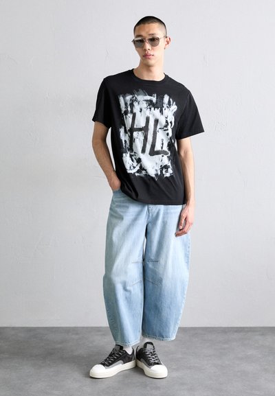 Fekete póló fehér absztrakt grafikával; világoskék, bő szabású farmernadrág; sötét tornacipők fehér talppal; karcsú napszemüveg.