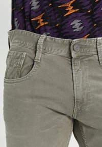 Jasnooliwkowe spodnie wykonane z denimu. Posiadają pięć kieszeni, zapięcie na guzik oraz widoczne przeszycia.