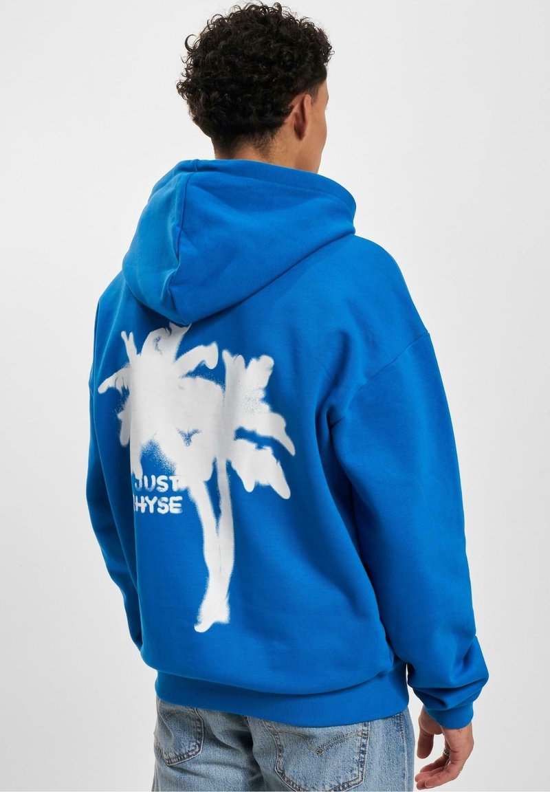 Personne portant un sweat à capuche bleu vif avec un motif palmier blanc et un texte partiellement visible dans le dos, associé à un jean bleu clair.