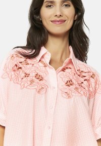 Camicia a quadri rosa con applicazioni in pizzo floreale su un lato, chiusura con bottoni e maniche corte, che offre un elemento di design testurizzato.