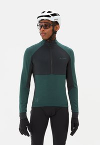 Maillot de cyclisme à manches longues en vert foncé et noir, avec une fermeture éclair à demi sur le devant, une texture douce et des accents réfléchissants. Porté avec des gants noirs.