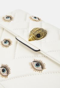 Vadderad vit läderklutch med guldögonmotiv på hårdvaran och pärlprydnader, med en klaffstängning och kontrasterande svart trim.
