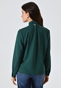 Blusa a maniche lunghe di colore verde scuro con colletto alto e piega sul retro. Caratterizzata da polsini con bottoni; texture liscia e design minimalista.