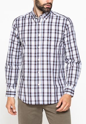 Uomo che indossa una camicia a quadri a maniche lunghe bianca, marrone e blu con pantaloni beige, su sfondo bianco semplice.