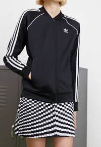 Person som bär en svart Adidas zip-up-jacka med vita ränder och en svart-vit rutad kjol, stående nära grå lockers.