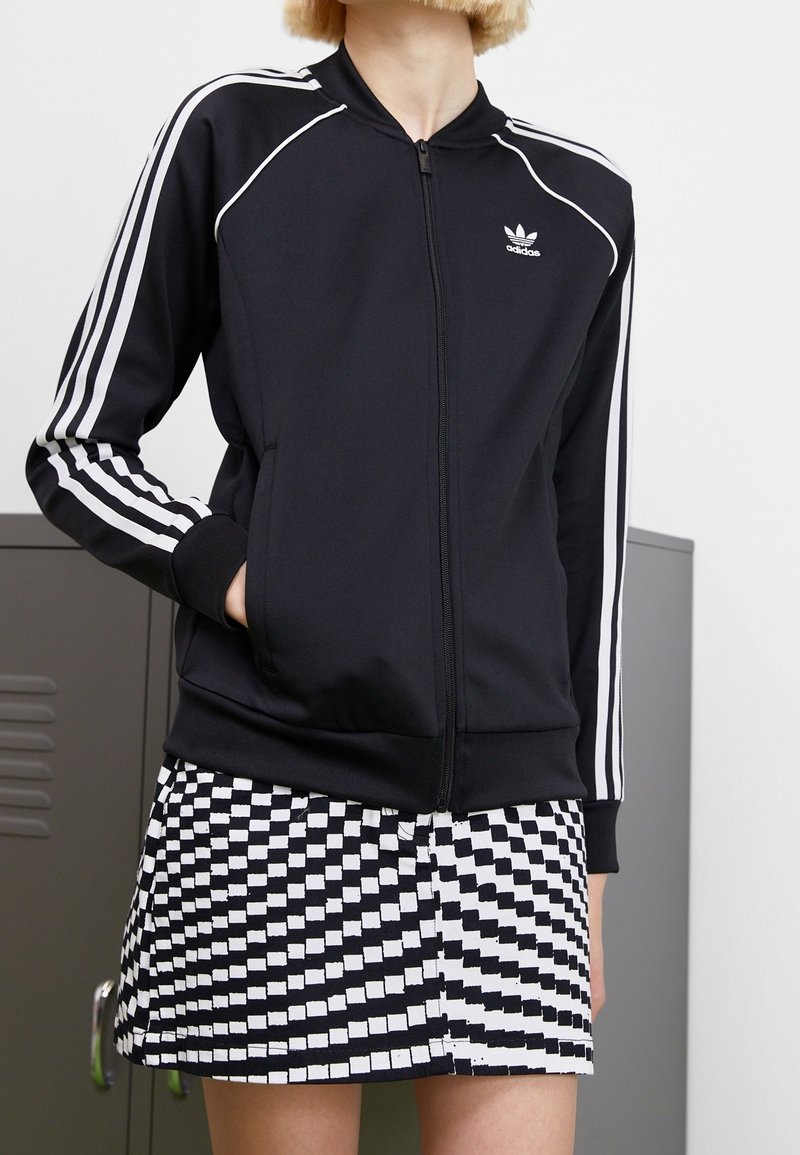 Person som bär en svart Adidas zip-up-jacka med vita ränder och en svart-vit rutad kjol, stående nära grå lockers.