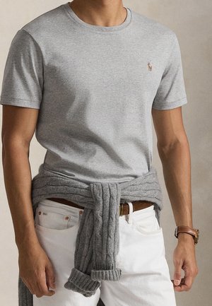 Homme portant un t-shirt gris clair avec un petit logo brodé, un pantalon blanc, une ceinture marron et un pull gris noué autour de la taille.