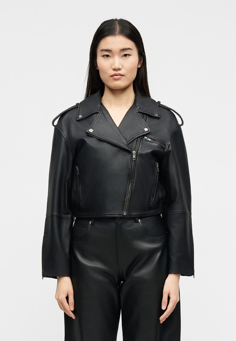 Νεαρή γυναίκα με μακριά σκούρα μαλλιά, που φοράει ένα μαύρο κομμένο δερμάτινο μπουφάν μοτοσικλετιστή και δερμάτινο παντελόνι με ψηλή μέση, μπροστά σε απλό φόντο.