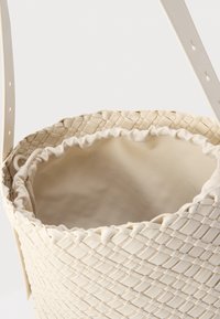 Sac tissé couleur crème, doté d'une surface extérieure texturée, d'une doublure intérieure lisse, et d'une bandoulière réglable avec des trous perforés pour personnaliser la longueur.