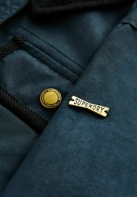 Närbild av mörk tyg med två gyllene metallmärken; ett runt med texten "Superdry Expedition Co." och ett rektangulärt med texten "Superdry".