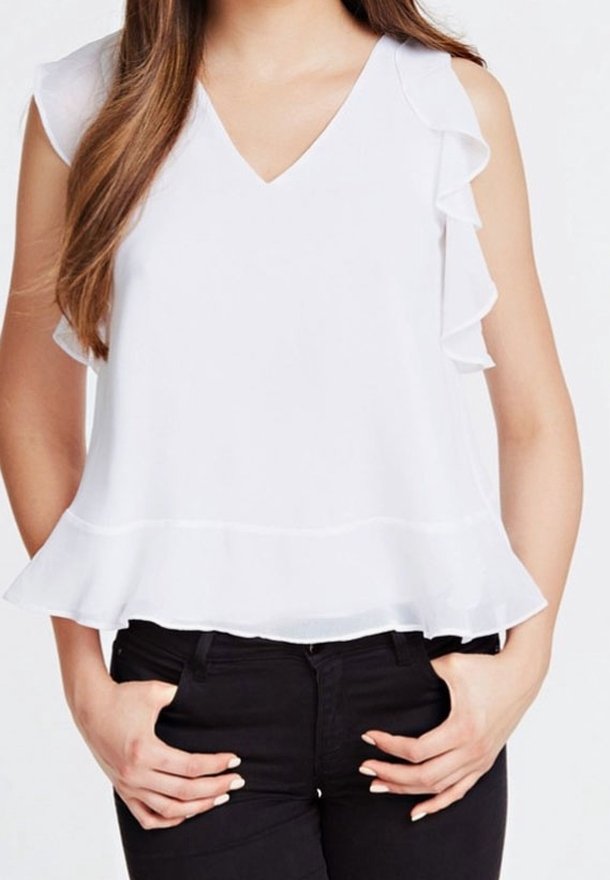 Femme portant un chemisier blanc sans manches à col en V avec des bordures à volants, associé à un pantalon noir, mains reposant dans les poches avant.