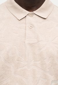 Μπεζ υφασμάτινο πόλο με δύο κουμπιά κλειστά, φορεμένο από ένα άτομο με σκουρόχρωμο δέρμα.