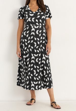 Femme portant une robe midi à motif géométrique noir et blanc, manches courtes, et des sandales noires, debout devant un fond uni.