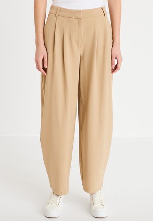 Osoba ubrana w beżowe spodnie o szerokich nogawkach i białe trampki, stojąca na tle jednolitego białego tła.