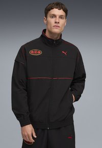 Ung mand iført en sort Puma tracksuit med røde detaljer og Sonic the Hedgehog-logo på jakken og bukserne, står med hånden i lommen.