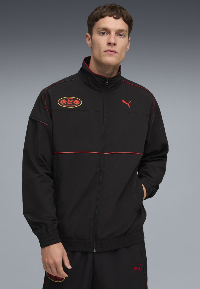 Ung mand iført en sort Puma tracksuit med røde detaljer og Sonic the Hedgehog-logo på jakken og bukserne, står med hånden i lommen.