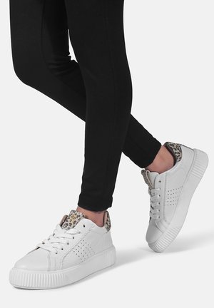 Fødder iført hvide sneakers med leopardprint på tungen og hælen, kombineret med sorte tætsiddende bukser mod en ensfarvet baggrund.