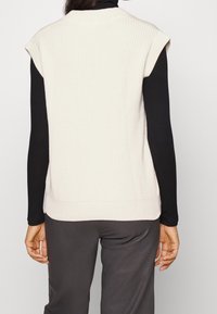 Helles beige, gerippte ärmellose Bluse mit rundem Halsausschnitt, getragen über einem schwarzen Langarmshirt. Verfügt über einen geraden Saum und eine strukturierte Stoffoberfläche.