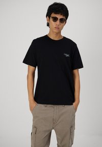 Svart bomulls t-skjorte med korte ermer, med en liten blå brodert logo på brystet. Sammen med lysebrune cargo-bukser.