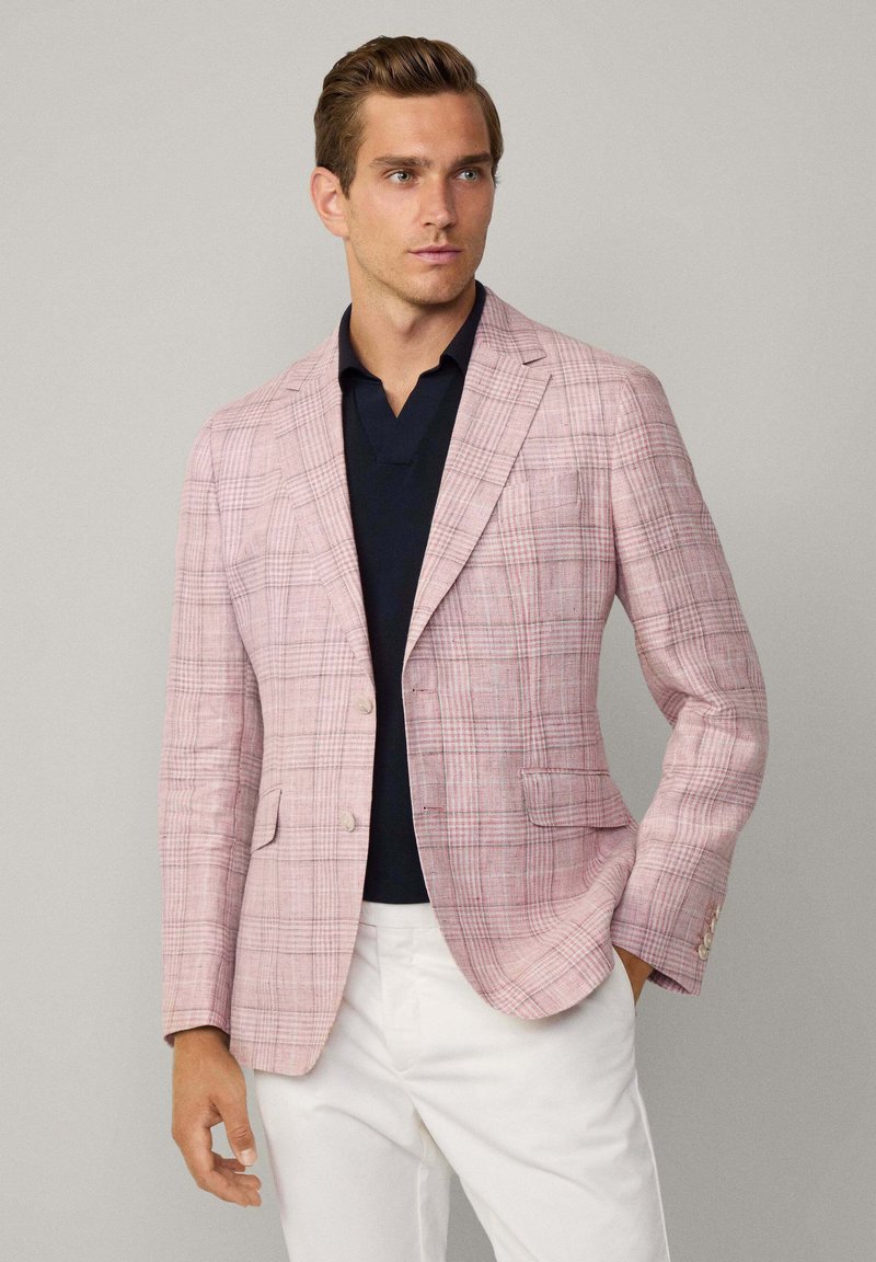 Blazer à carreaux rose clair en tissu texturé ; doté de deux boutons, de revers notch et de deux poches avant. Porté sur un polo noir.
