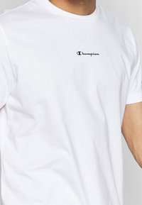 Hvid bomulds-T-shirt med korte ærmer, rund halsudskæring og et lille sort "Champion" logo broderet på venstre bryst.
