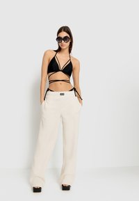 Zwarte bikini-top met dunne banden, in combinatie met crèmekleurige wijde broek van zachte stof. Model draagt een oversized zwart zonnebril.