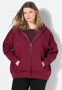 Sudadera borgoña con cremallera, con un bolsillo frontal, corte holgado y capucha con cordón; combinada con una blusa negra con estampado debajo.