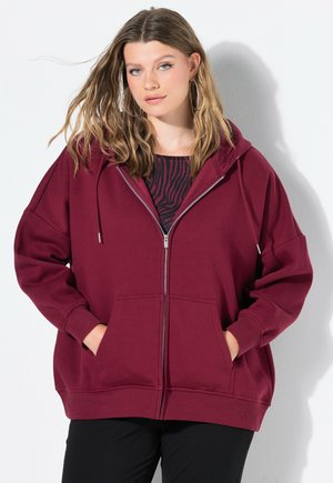 Haut à capuche zippé bourgogne avec une poche frontale, coupe ample et capuche à cordon ; associé à un haut noir à motifs en dessous.