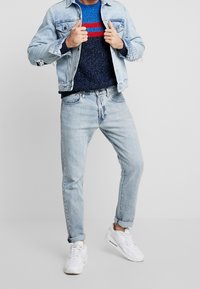 Jasnoniebieska kurtka dżinsowa nałożona na granatowo-niebieski sweter o teksturze z czerwonymi paskami, w zestawie z jasnymi dżinsami i białymi trampkami.