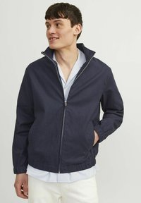 Tumed sinine kerge jakki, millel on kõrge krae, täispikki tõmblukk, elastsed mansetid ja hem, mis on valmistatud siledast kangast, millel on peen tekstuur.