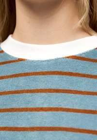 Κοντινό πλάνο ενός ατόμου που φοράει μπλε πουκάμισο από πετσέτα με οριζόντιες καφέ ρίγες και λευκό ριμπ ύφασμα στο λαιμό.