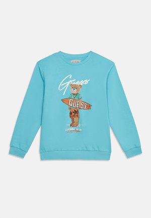 Vaaleansininen collegepaita, jossa on nallekarhu, joka pukeutunut petroolinväriseen paitaan ja oranssit shortsit, ja jonka kädessä on oranssi surffilauta, jossa on "GUESS"-teksti.