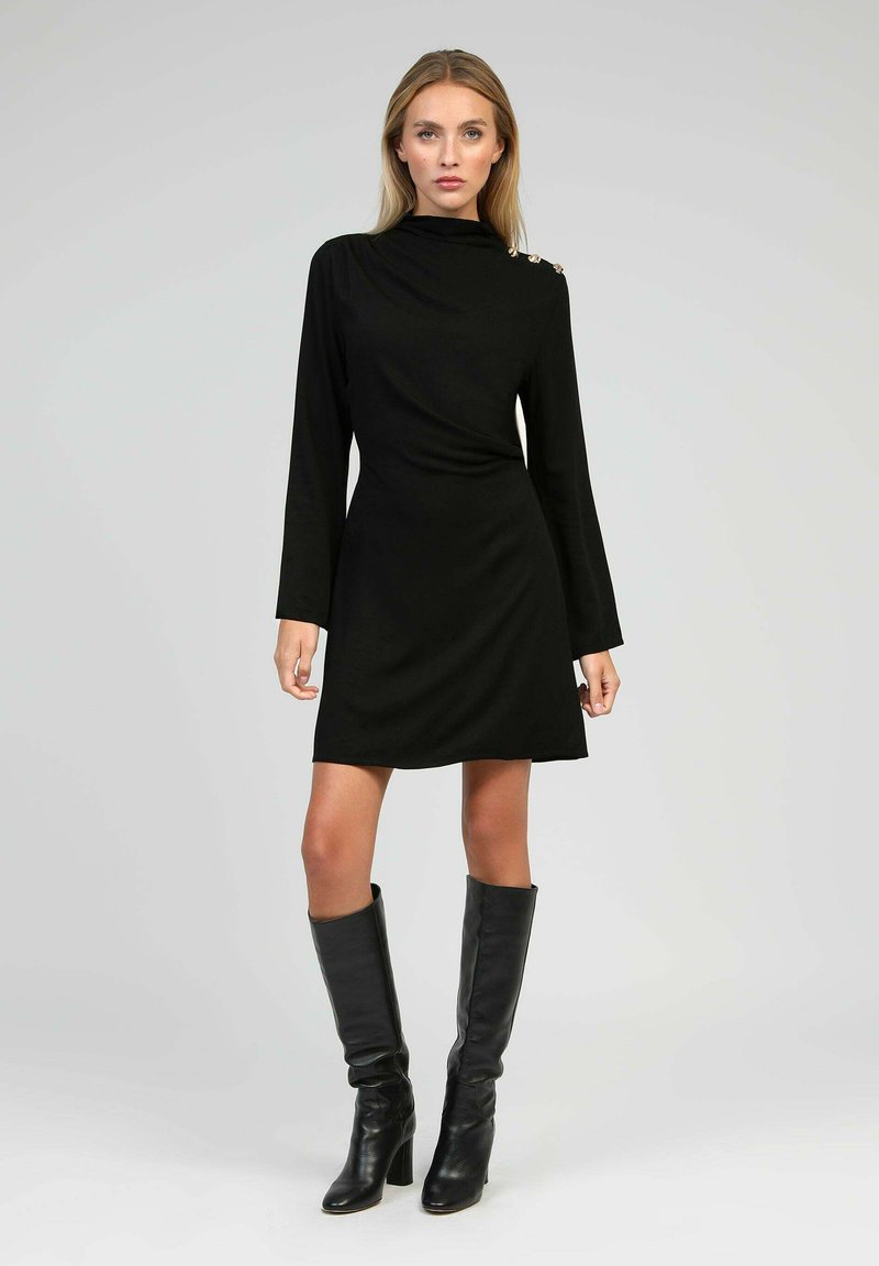 Femme portant une robe longue à manches noires avec des boutons dorés sur l'épaule, chaussée de bottes en cuir noir montantes, se tenant devant un fond gris clair.